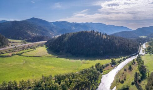 Aerial view of rolling green hills, forested slopes, and a winding road under a bright blue sky with scattered clouds.