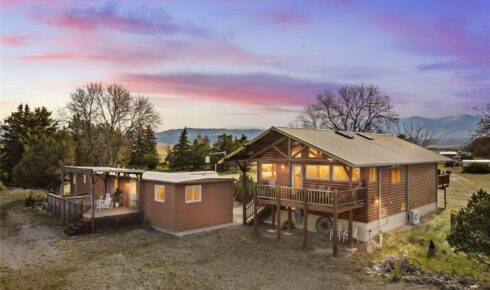 Rustic wooden cabin with a wraparound deck and a small shed, set on a dirt yard at sunset with mountains in the distance