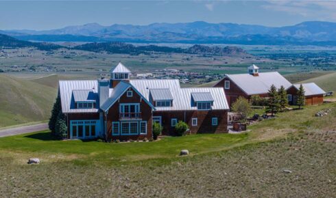 Large rustic wooden house with multiple wings and metal roofs on a grassy hillside, with distant mountains in the background.