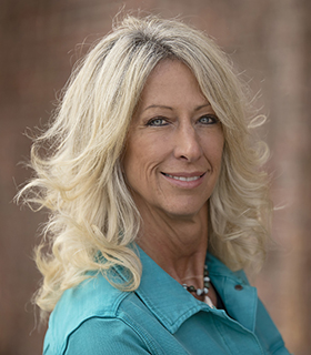 Portrait of a smiling blonde woman wearing a teal shirt outdoors, blurred brick background behind her.