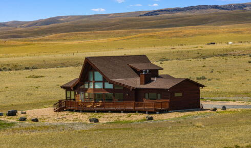 Wooden house with a wraparound deck in a wide grassy plain and rolling hills under a blue sky.
