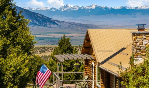 Log cabin with a tan metal roof and stone chimney, an American flag in the yard, and snow-capped mountains in the distance behind.