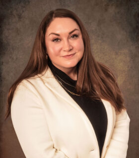 Head-and-shoulders portrait of a smiling woman with long brown hair, wearing a cream blazer over a black top, against a textured brown backdrop.
