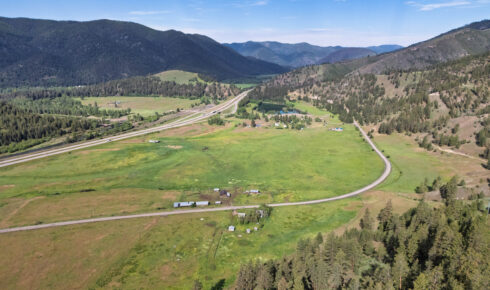 Aerial view of a rural valley with winding roads, green fields, and forested mountains under a blue sky.